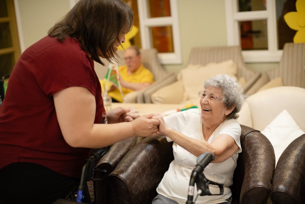 An older adult receives gentle assistance with grooming and dressing in a bright, comfortable bedroom.
