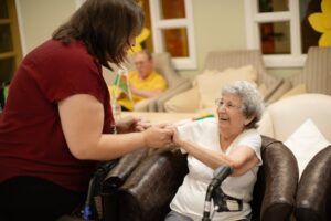 An older adult receives gentle assistance with grooming and dressing in a bright, comfortable bedroom.