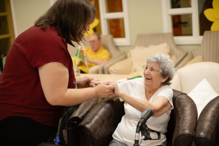 An older adult receives gentle assistance with grooming and dressing in a bright, comfortable bedroom.
