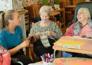 Smiling older adult and a caregiver sitting together at a kitchen table sharing conversation and coffee.