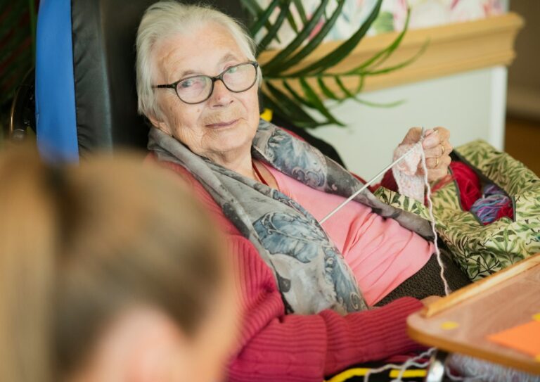 An older adult and companion share conversation at a kitchen table beside family photographs.